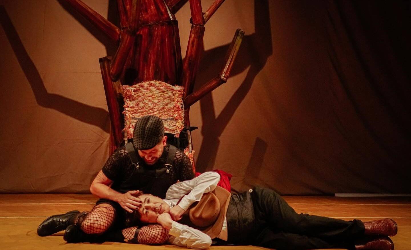 Dois atores em trajes de palco, um sentado e outro deitado no chão, em uma cena teatral com um cenário rústico.