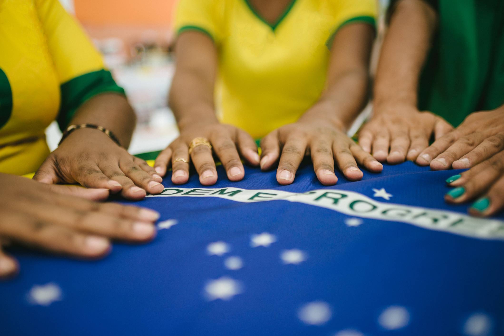 Mãos de pessoas com camisas amarela e verde repousando sobre a bandeira do Brasil, com parte da inscrição e estrelas visíveis.