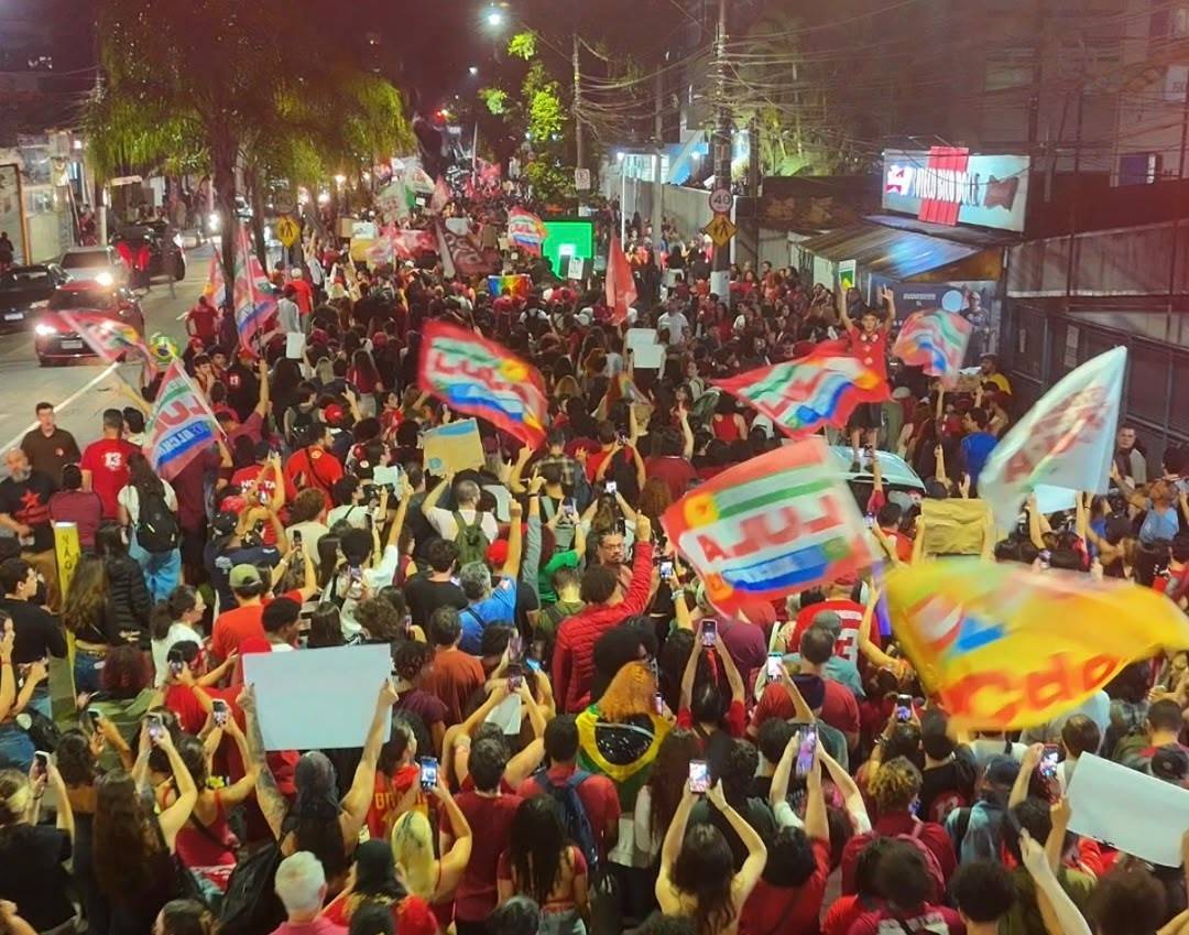 Multidão em manifestação política noturna com muitas pessoas vestindo vermelho, agitando bandeiras e segurando celulares
