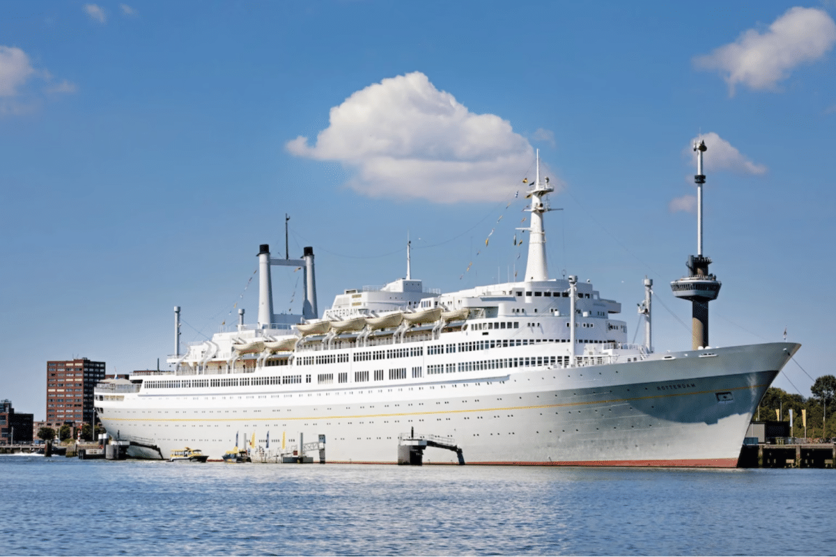 Navio de cruzeiro branco 'Rotterdam' atracado em águas azuis calmas, sob céu claro com nuvens e torre de observação ao fundo.