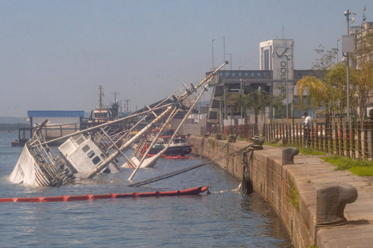 Barco grande parcialmente submerso e inclinado em um porto, com barreira de contenção de óleo laranja na água. Edifício 'Parque Valongo' ao fundo.