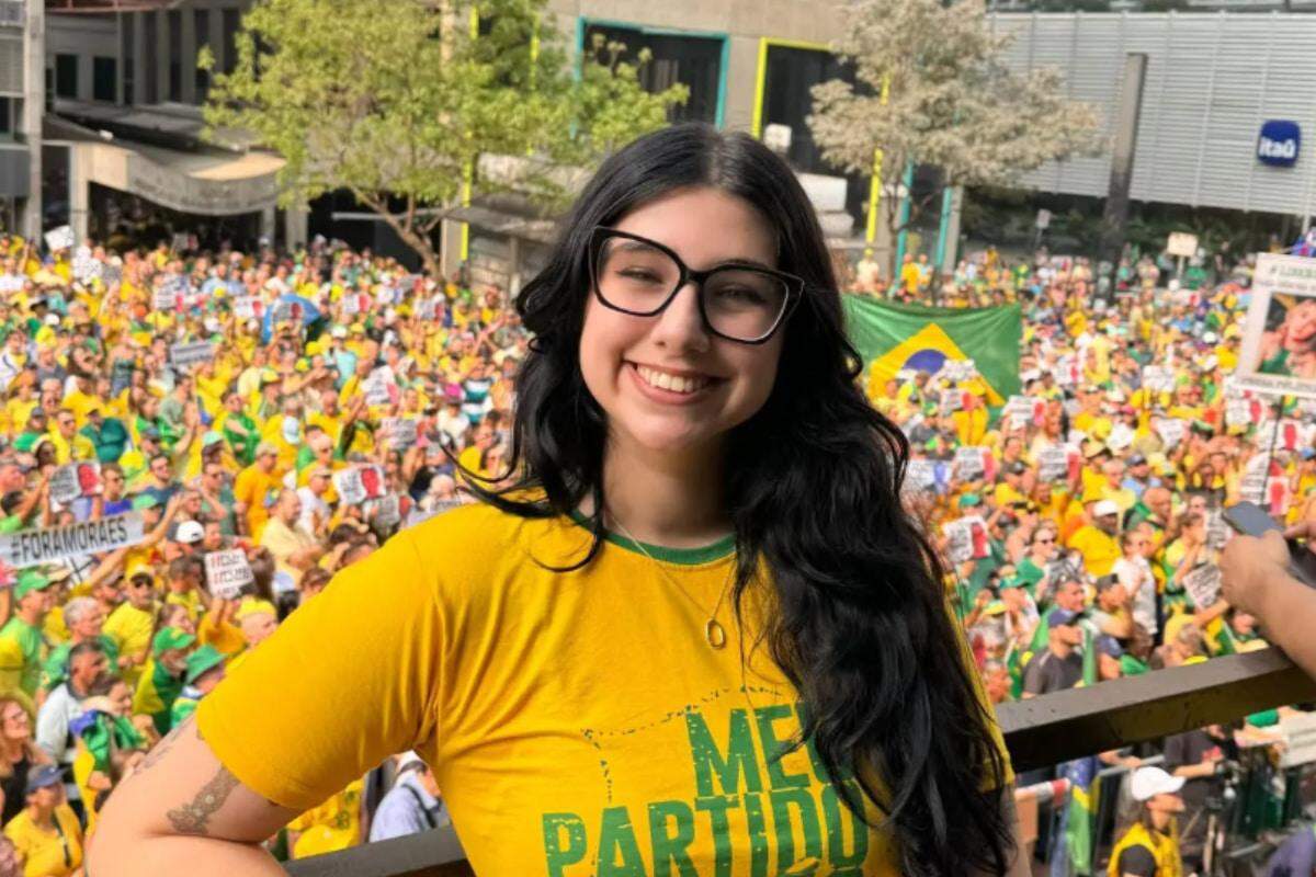 Young woman in yellow shirt smiling, glasses, in front of a large crowd wearing yellow/green, holding Brazilian flags and protest signs.