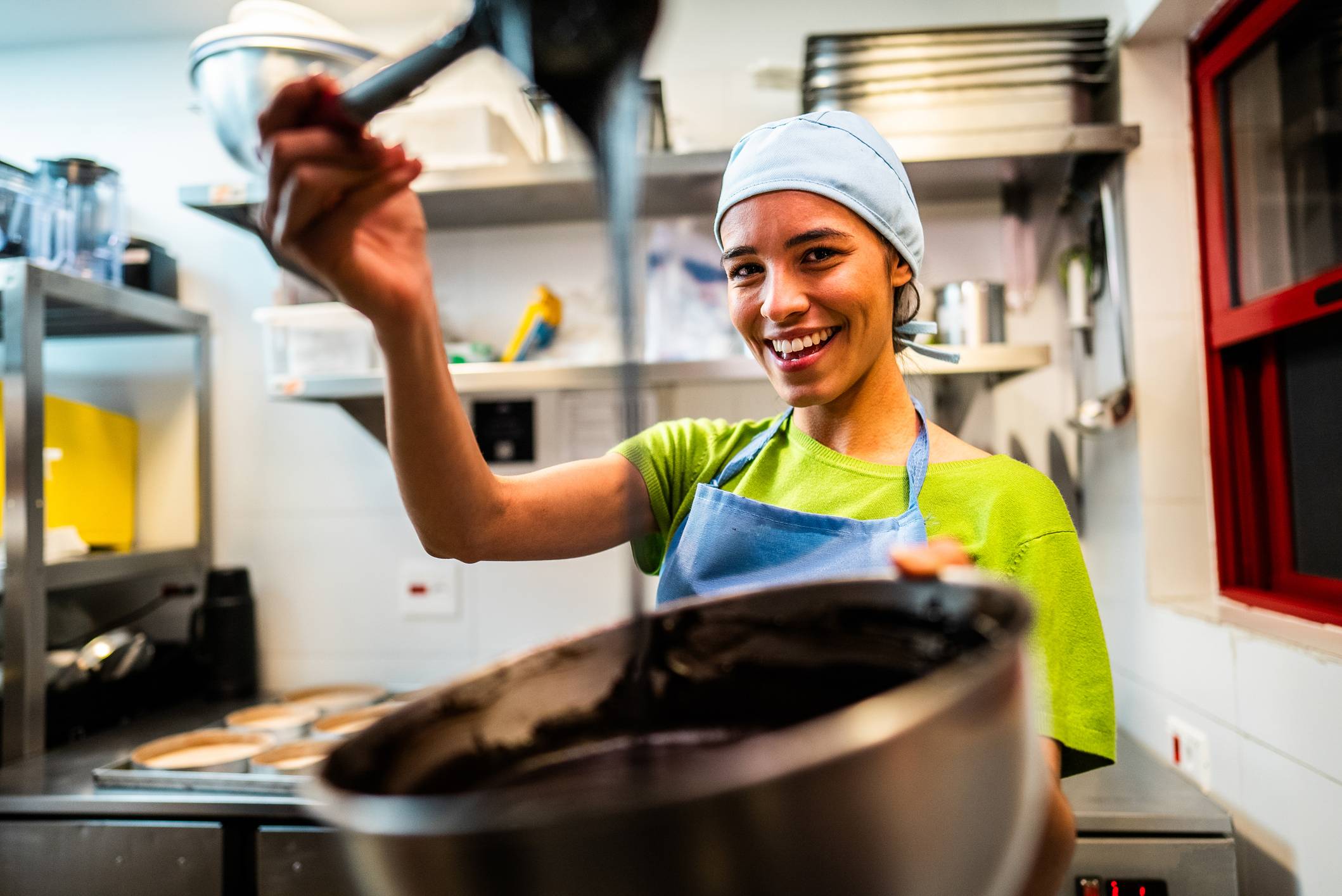 Mulher jovem sorrindo em cozinha profissional, despejando massa escura de uma concha em tigela de metal.