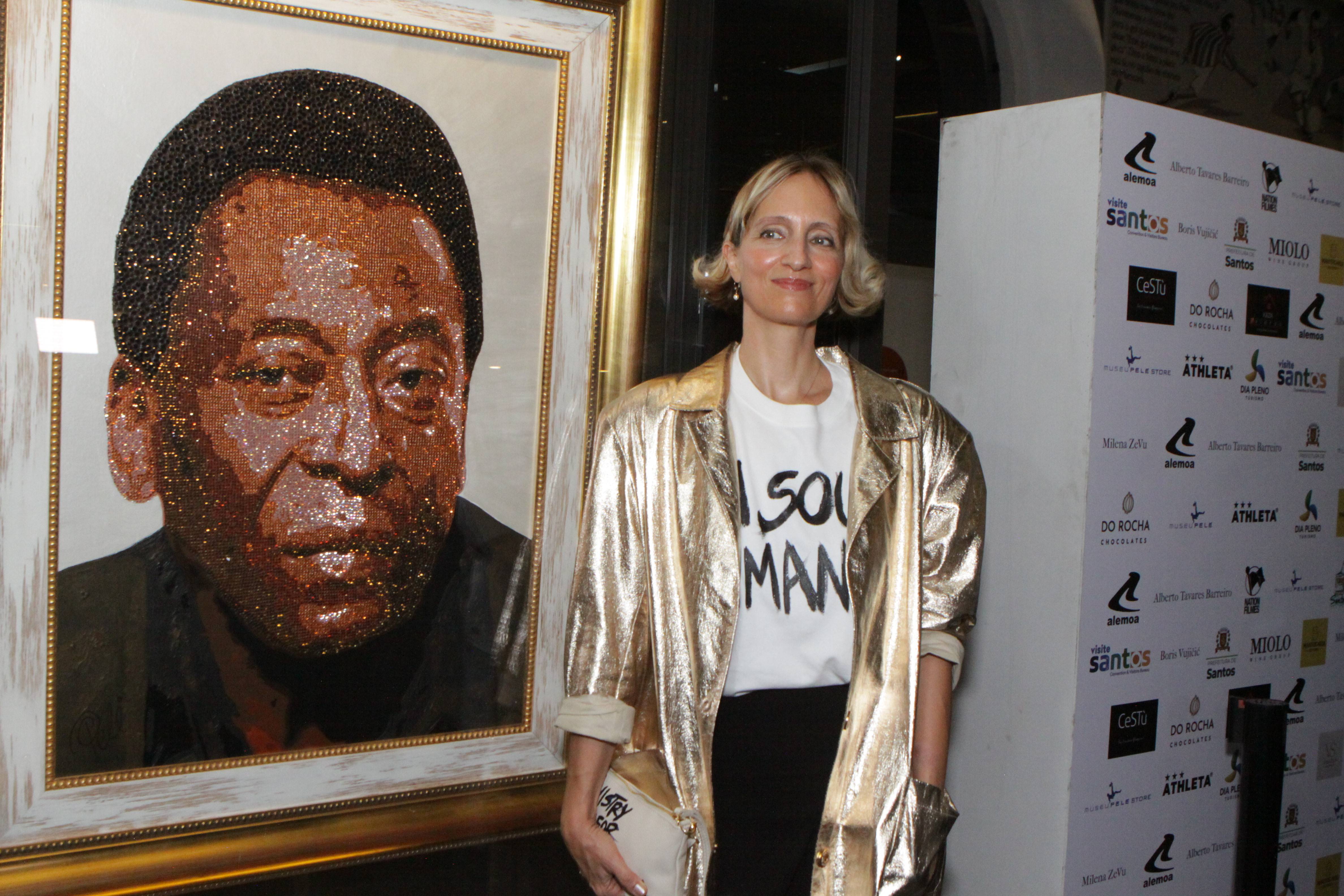 Mulher de jaqueta dourada ao lado de um retrato brilhante de Pelé feito com lantejoulas em evento com patrocinadores.