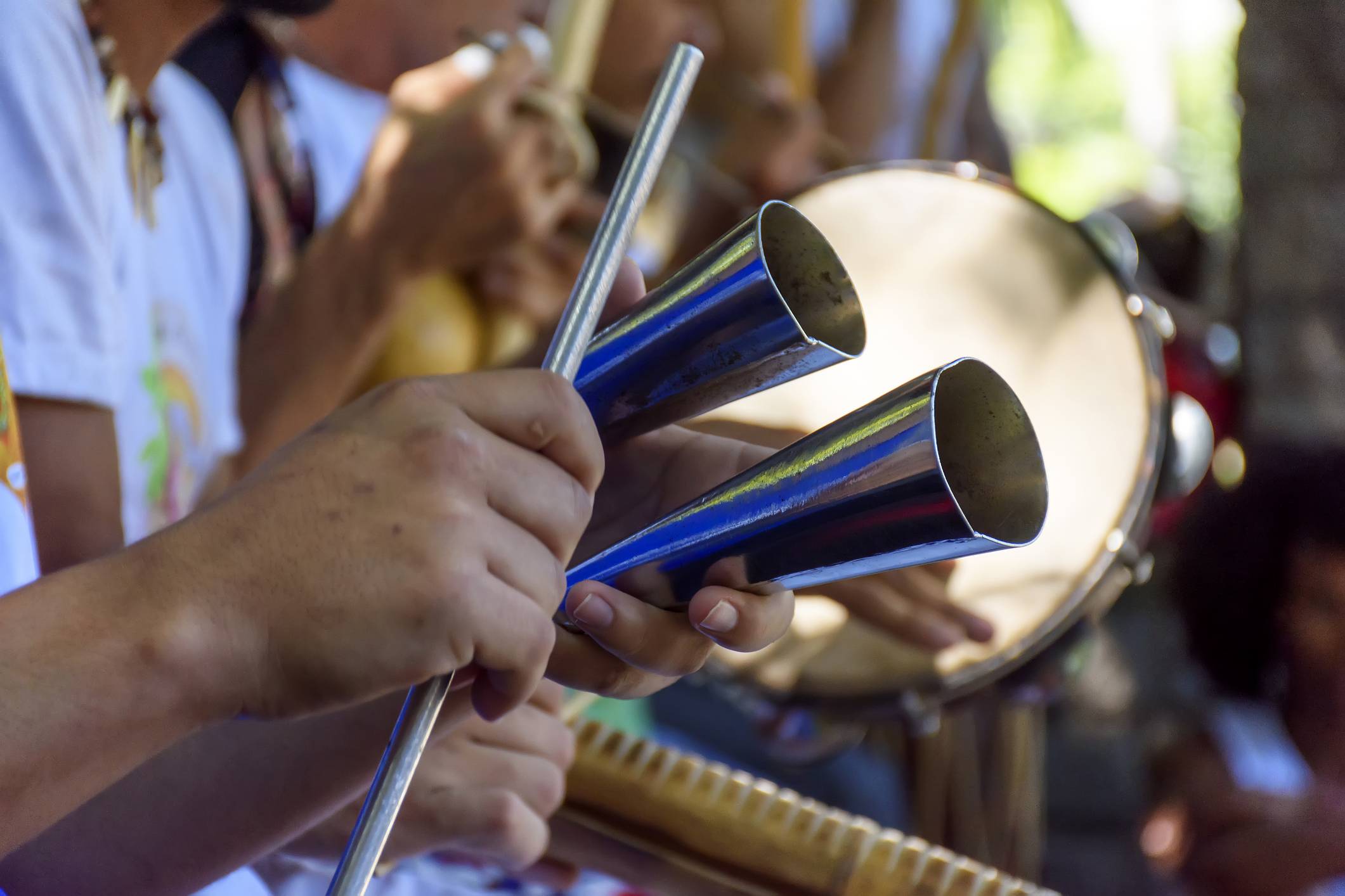Mãos tocam agogô duplo com baqueta metálica, com outros percussionistas e um tambor ao fundo em performance musical.