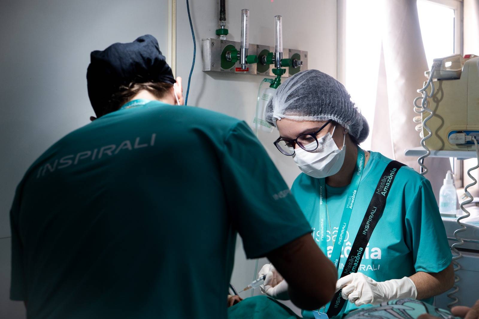 Profissionais de saúde em scrubs verdes, um de costas e uma mulher de óculos, máscara e luvas, segurando uma seringa em ambiente clínico.