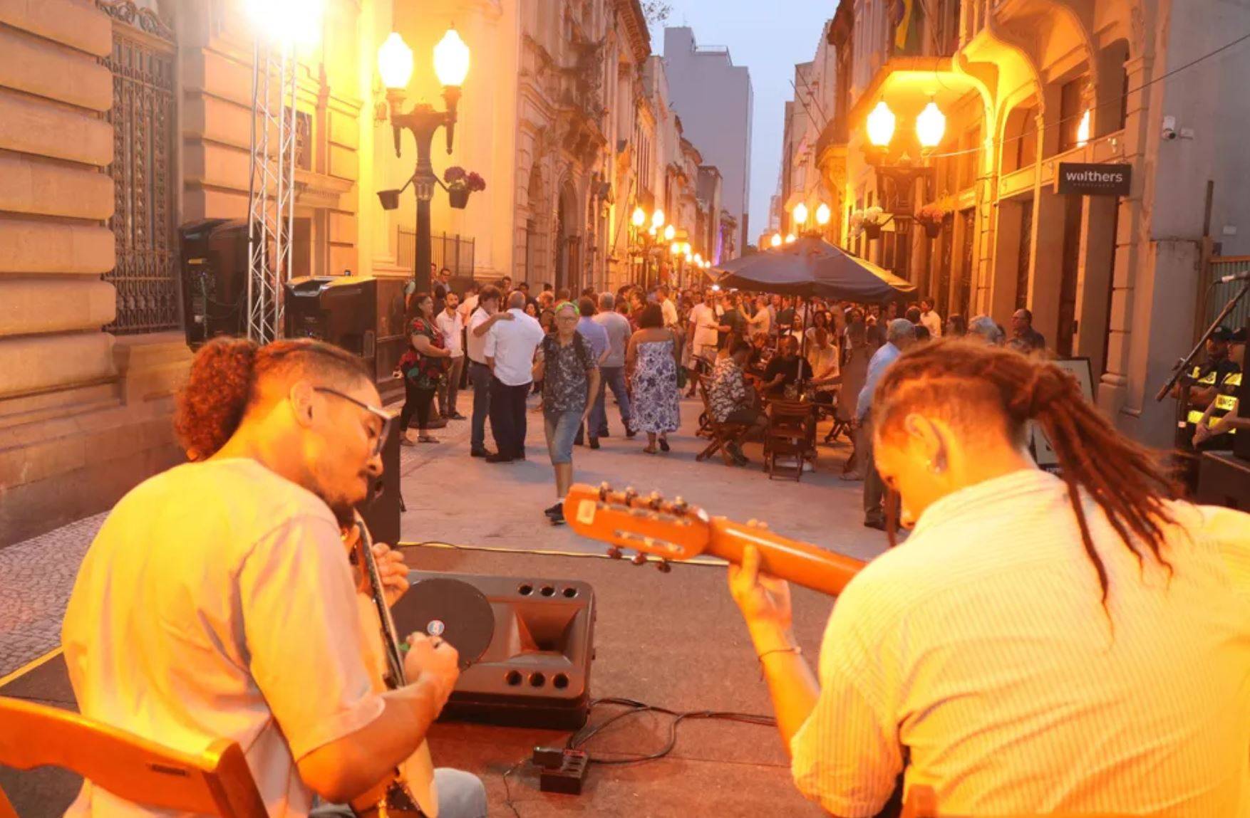 Dois músicos se apresentam ao vivo ao ar livre à noite para um público em uma rua iluminada por luzes quentes.