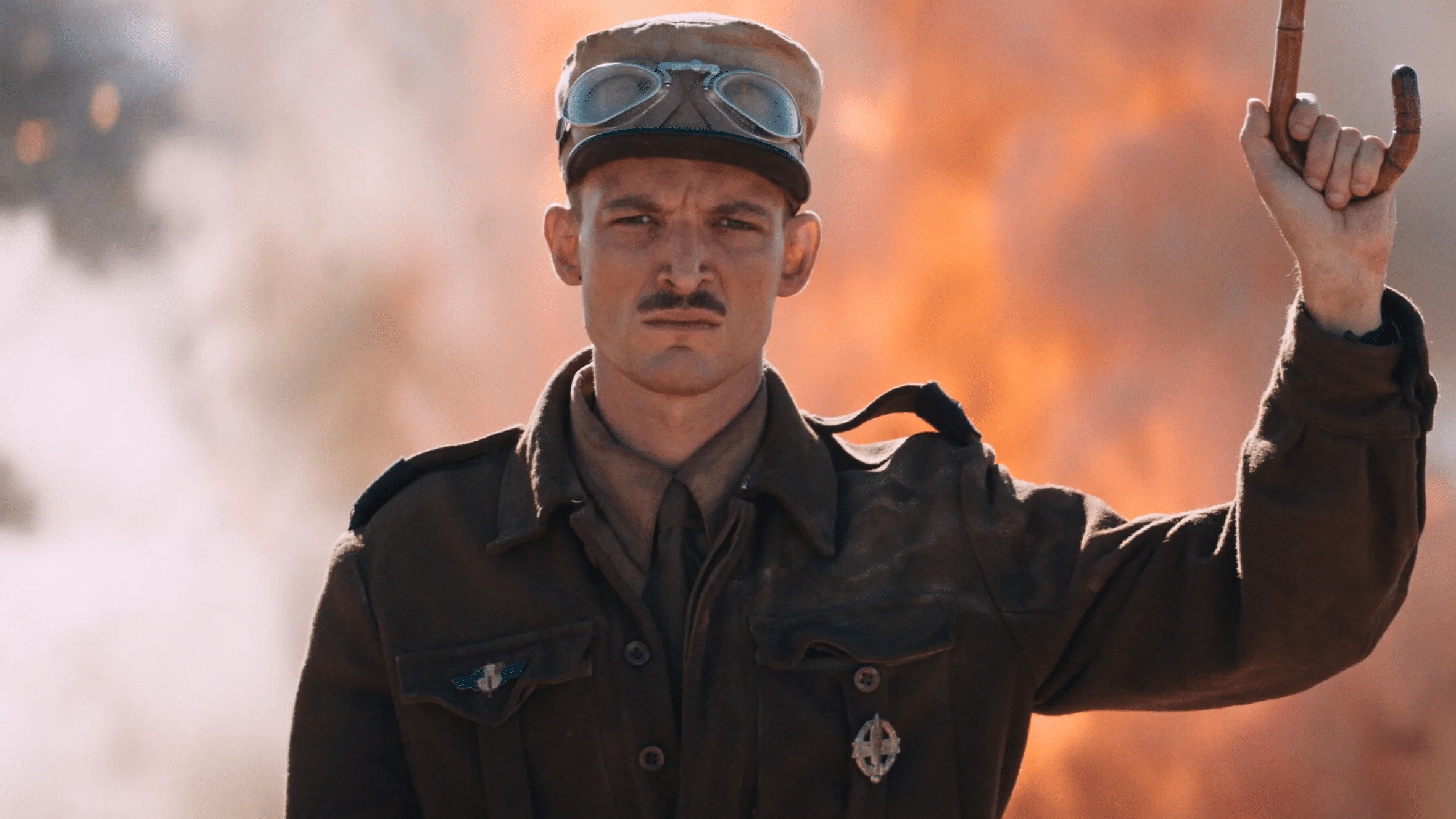 Homem de uniforme militar vintage, bigode e óculos na touca, ergue bengala em cenário de fumaça e fogo.