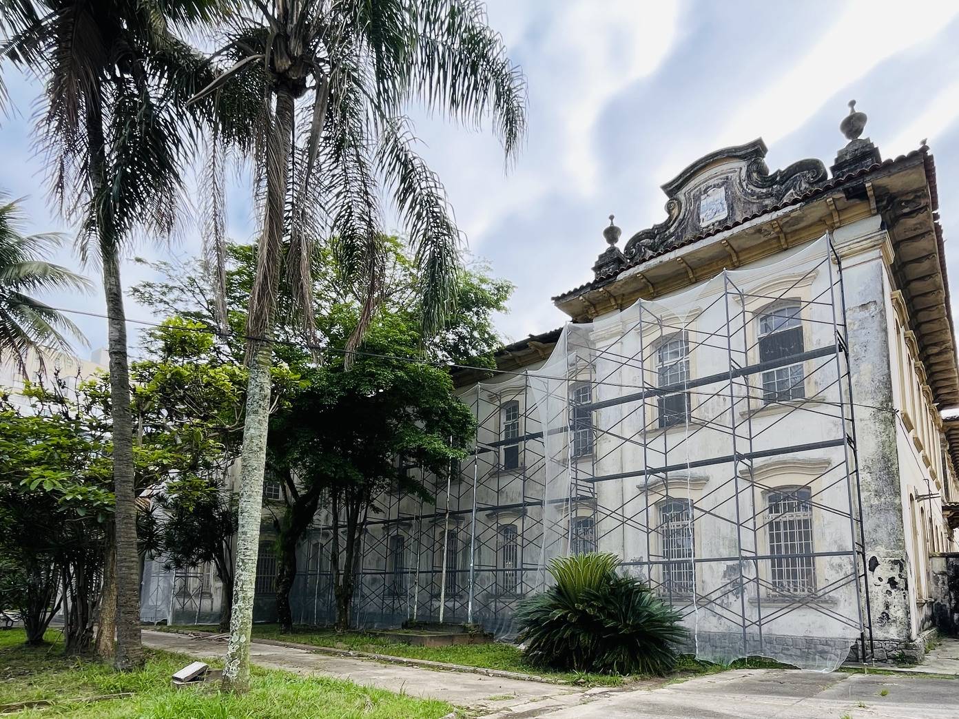 Entrada de edifício antigo com porta de madeira entreaberta, degraus de concreto, paredes descascadas e janelas decorativas amarelas.