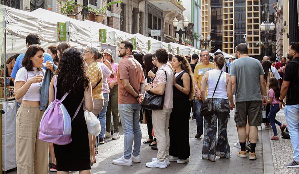 Multidão de pessoas caminhando e interagindo em rua movimentada com tendas brancas de mercado e edifícios ao fundo.
