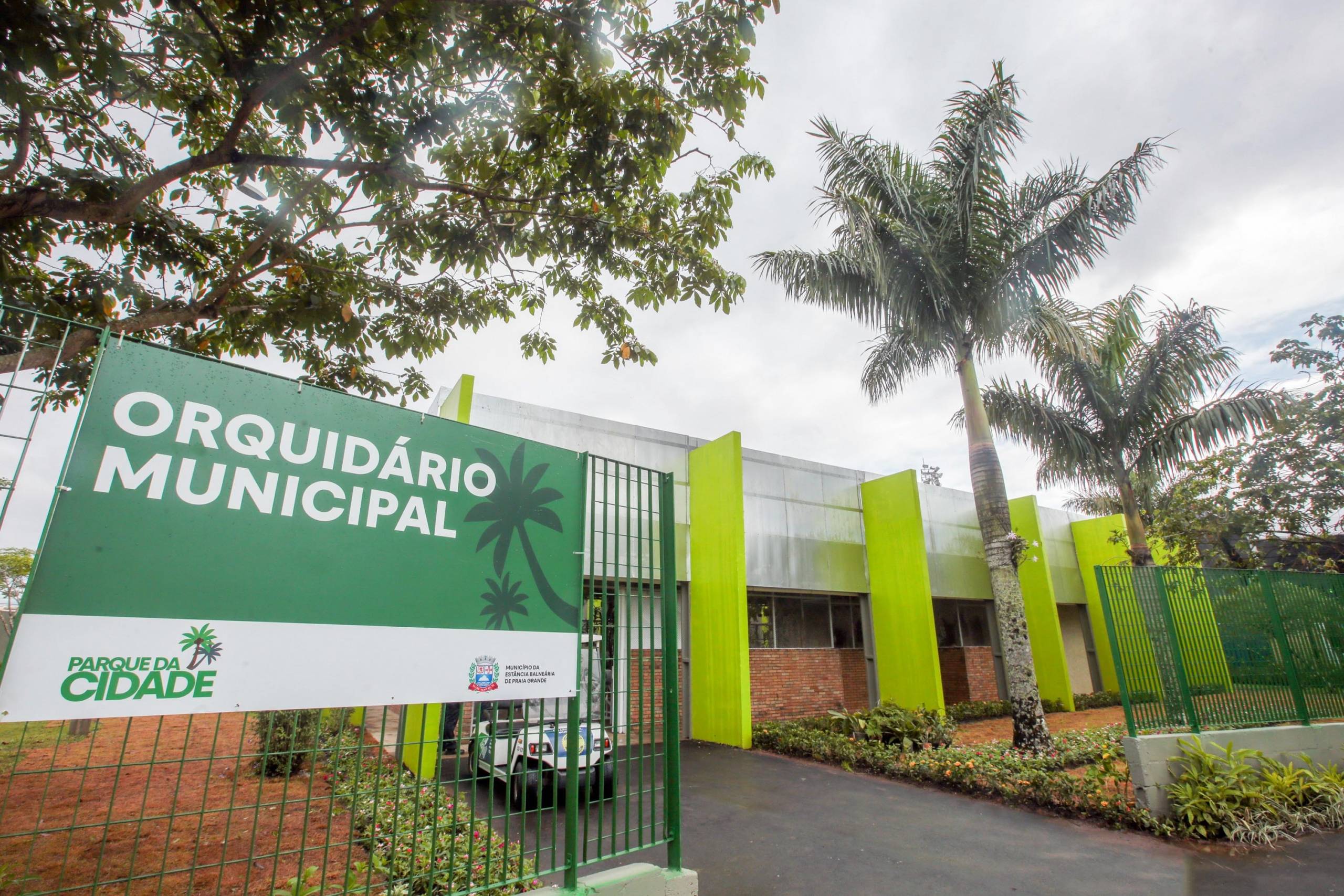 Placa verde e branca com os dizeres 'Orquidário Municipal' e 'Parque da Cidade' em frente a um edifício moderno com detalhes verdes e palmeiras.