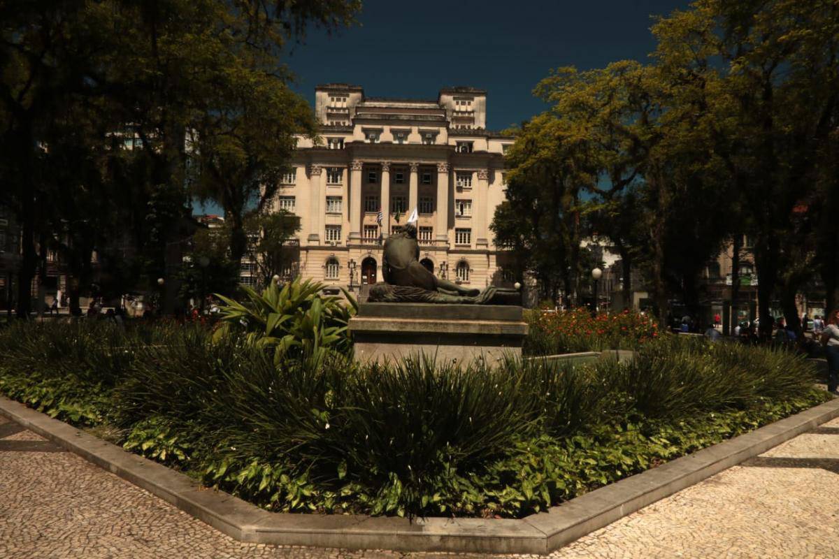 Praça com estátua de bronze de figura reclinada e edifício neoclássico de vários andares ao fundo, cercado por árvores.
