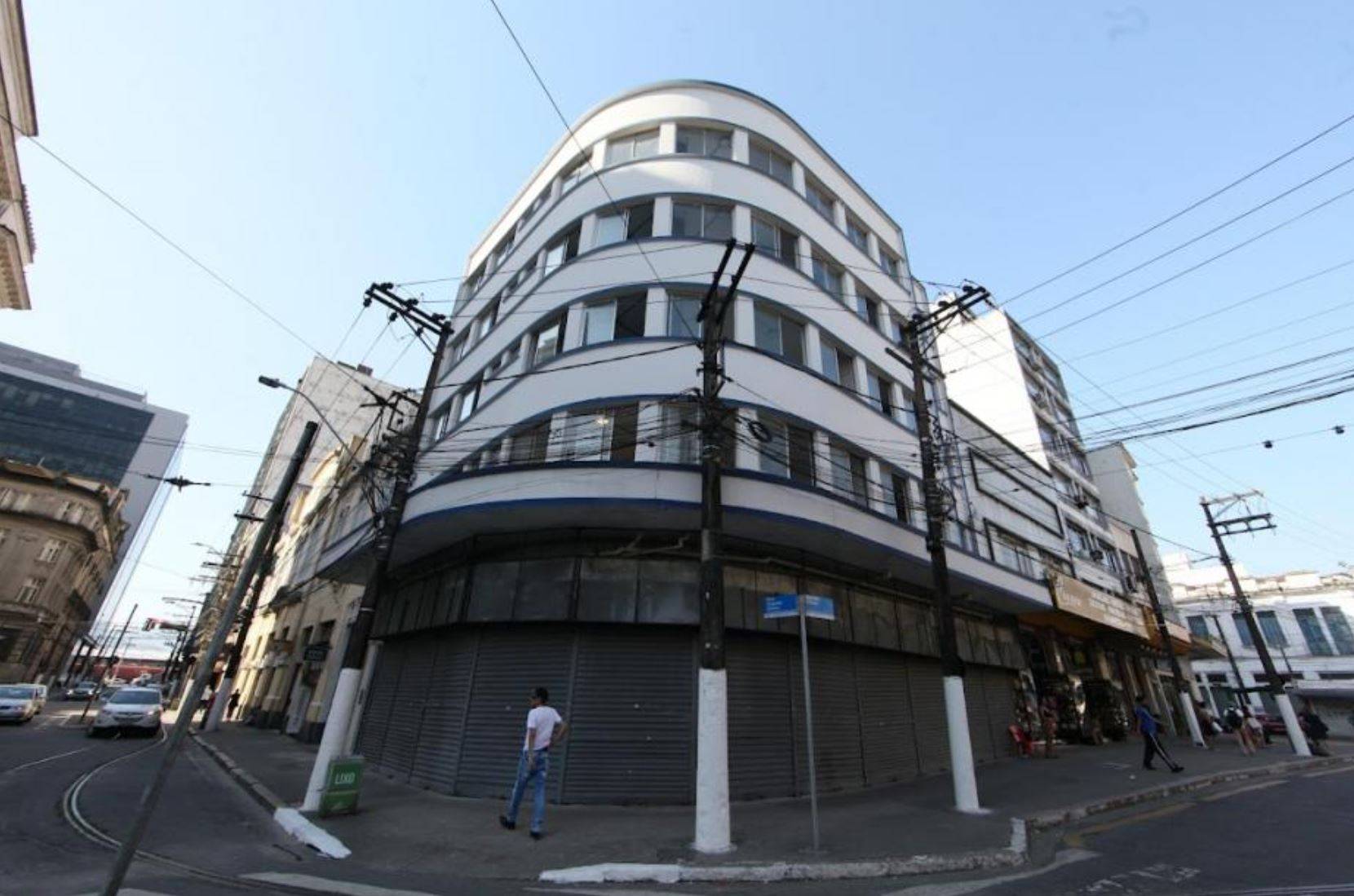 Edifício branco curvo com janelas e persianas fechadas em esquina urbana, postes com fiação exposta e um homem caminhando.