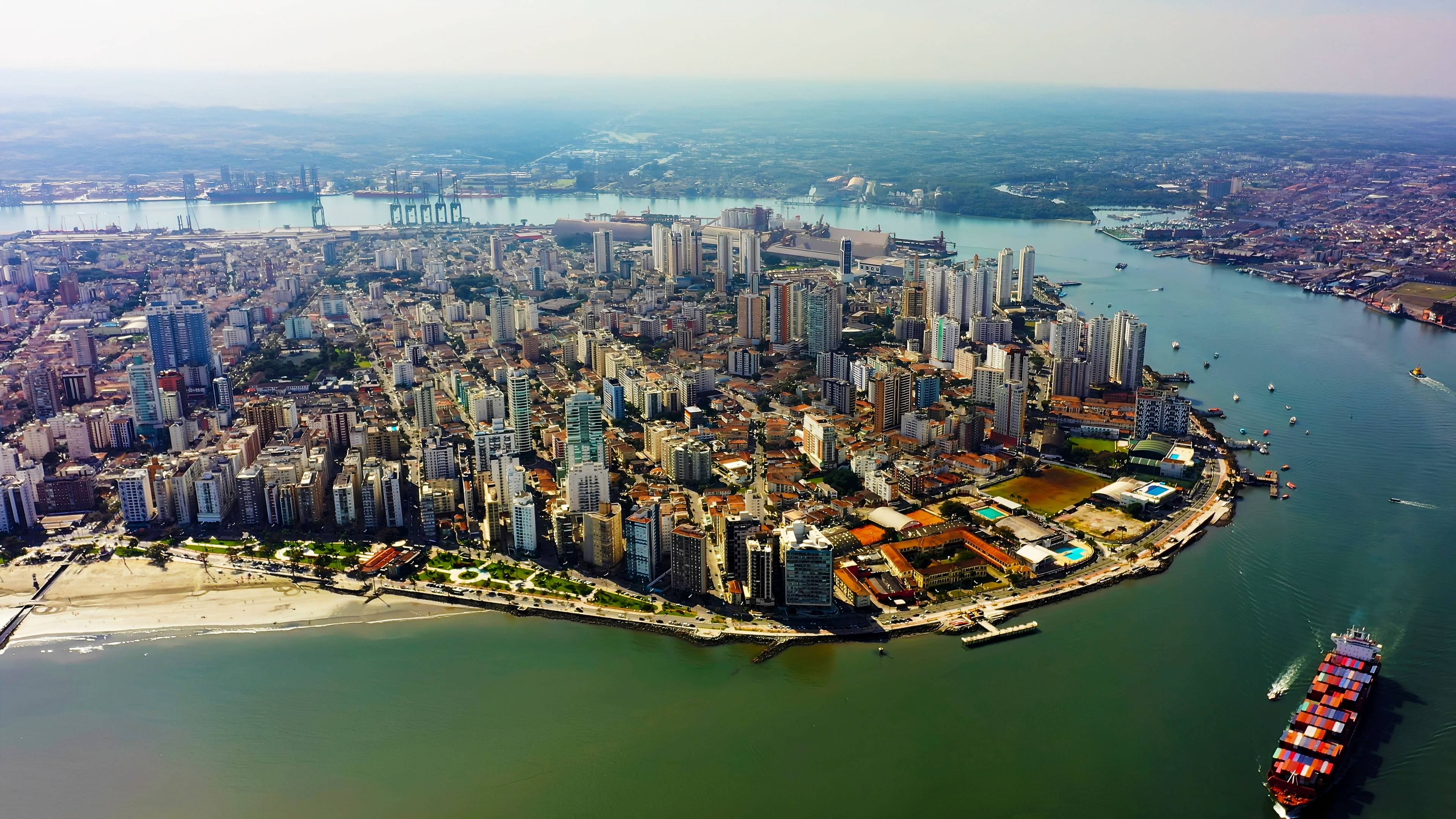 Vista aérea de Santos, SP, com navio cargueiro, praia, prédios e o porto ao fundo, sob céu claro.